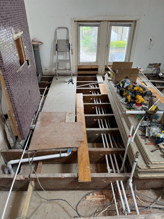 Opened floor showing joists and plastic pipework with ladder and tools beside a tiled wall