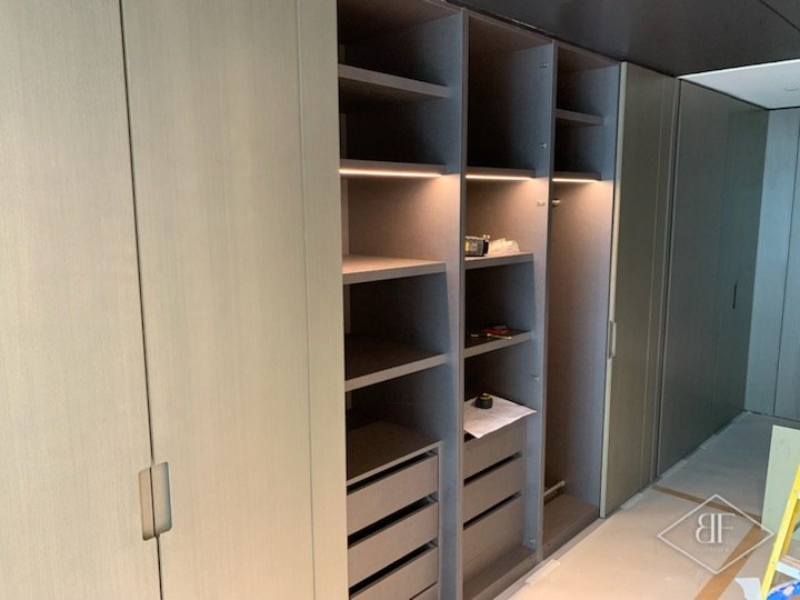 Fitted wardrobes with open compartments showing lit shelves and drawers, doors still being fitted