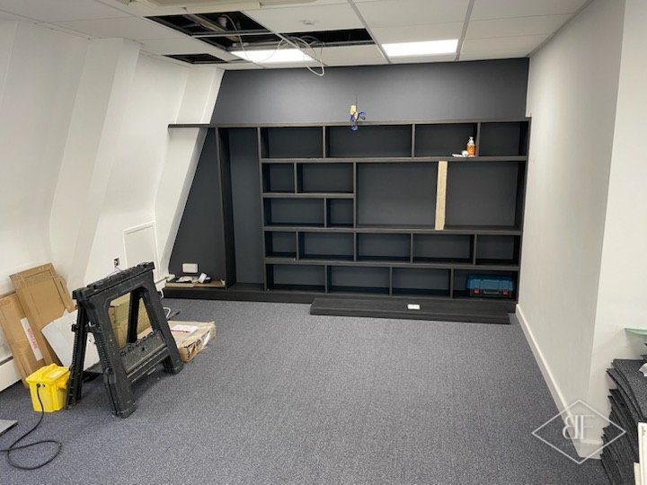 Dark shelving wall nearing completion in an office, with trestle and packaging on the carpet