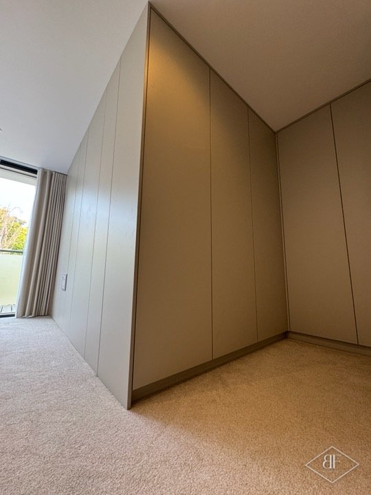 Angled handleless wardrobe doors beside a bedroom window with curtains, finished in a soft taupe