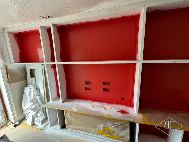 Red back panels being sprayed inside the white-primed shelving, with dust sheets protecting the room
