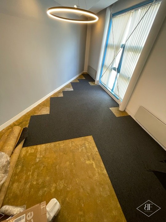 Office floor being covered with dark carpet tiles laid from the window end