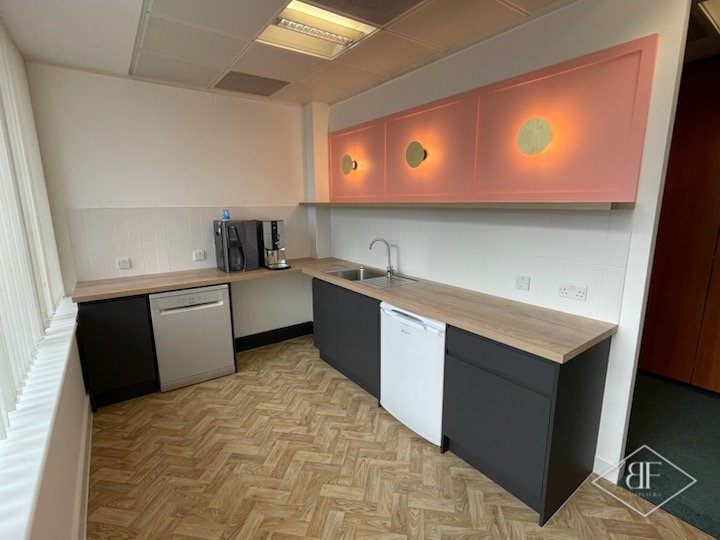 Office kitchenette with dark units, oak-effect worktop and illuminated coral wall cabinets