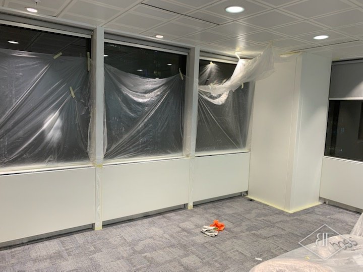 Window glazing covered in plastic sheeting while white perimeter panels are sprayed
