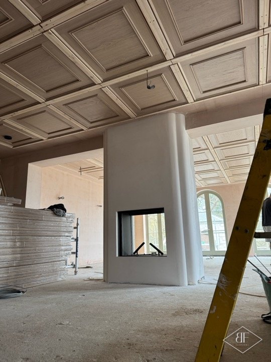 Large open room with coffered ceiling and central fireplace column awaiting flooring